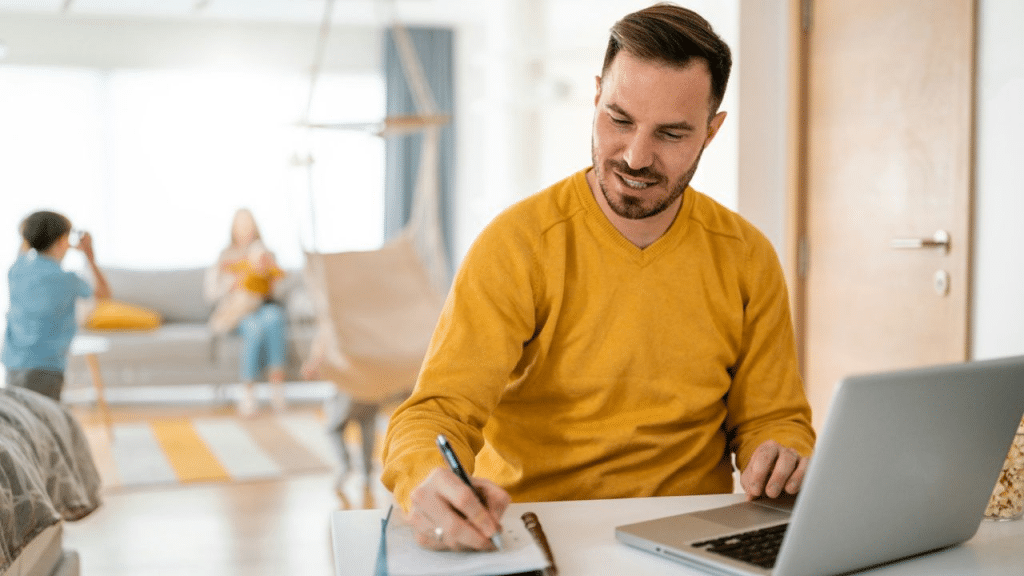 A man sits at a desk, looking at a laptop and writing in a notebook. People are blurry in the background.
