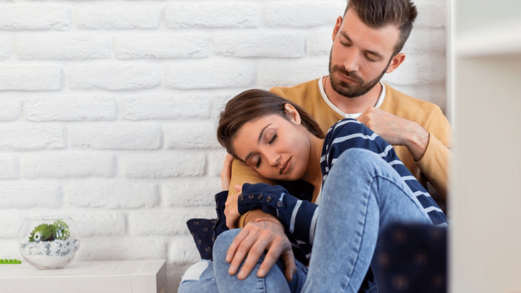 A man with a beard sits on a sofa, comforting a woman who is resting her head on his shoulder.