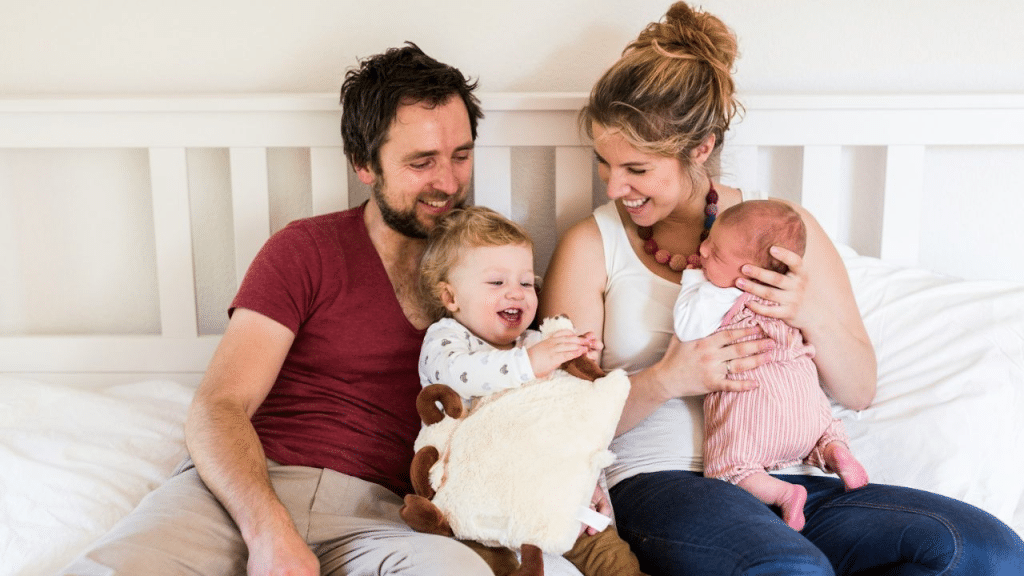 A happy couple sits on a bed with their baby and a toddler.