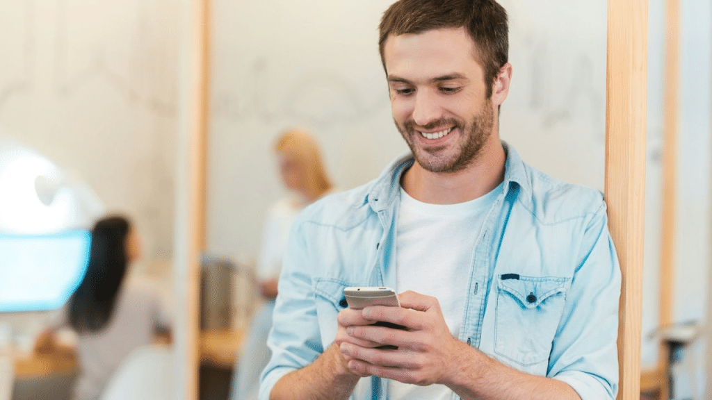 A smiling man looks down at his phone while leaning against a wooden wall.