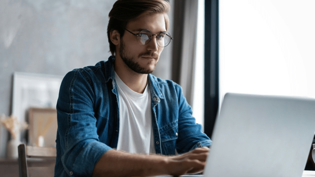 A man with glasses and a beard sits at a desk, looking at his laptop.