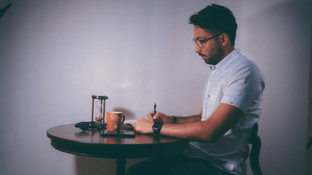 A man sits at a round table, writing in a notebook with a pen. An hourglass and a coffee mug are on the table.