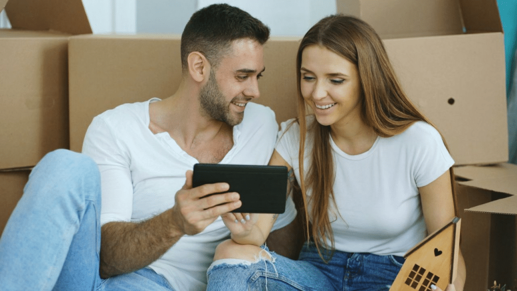 A smiling couple sits on the floor surrounded by boxes, looking at a tablet.