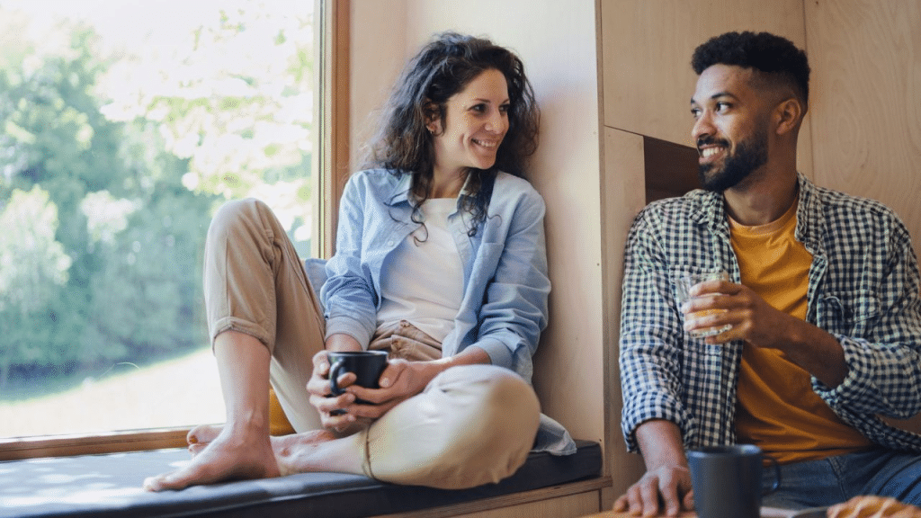 A happy couple sits together in a window nook, talking and holding drinks.