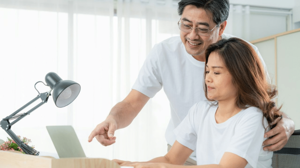 A happy couple in white shirts smiles while looking at a laptop together at a desk. The man is pointing at the screen.