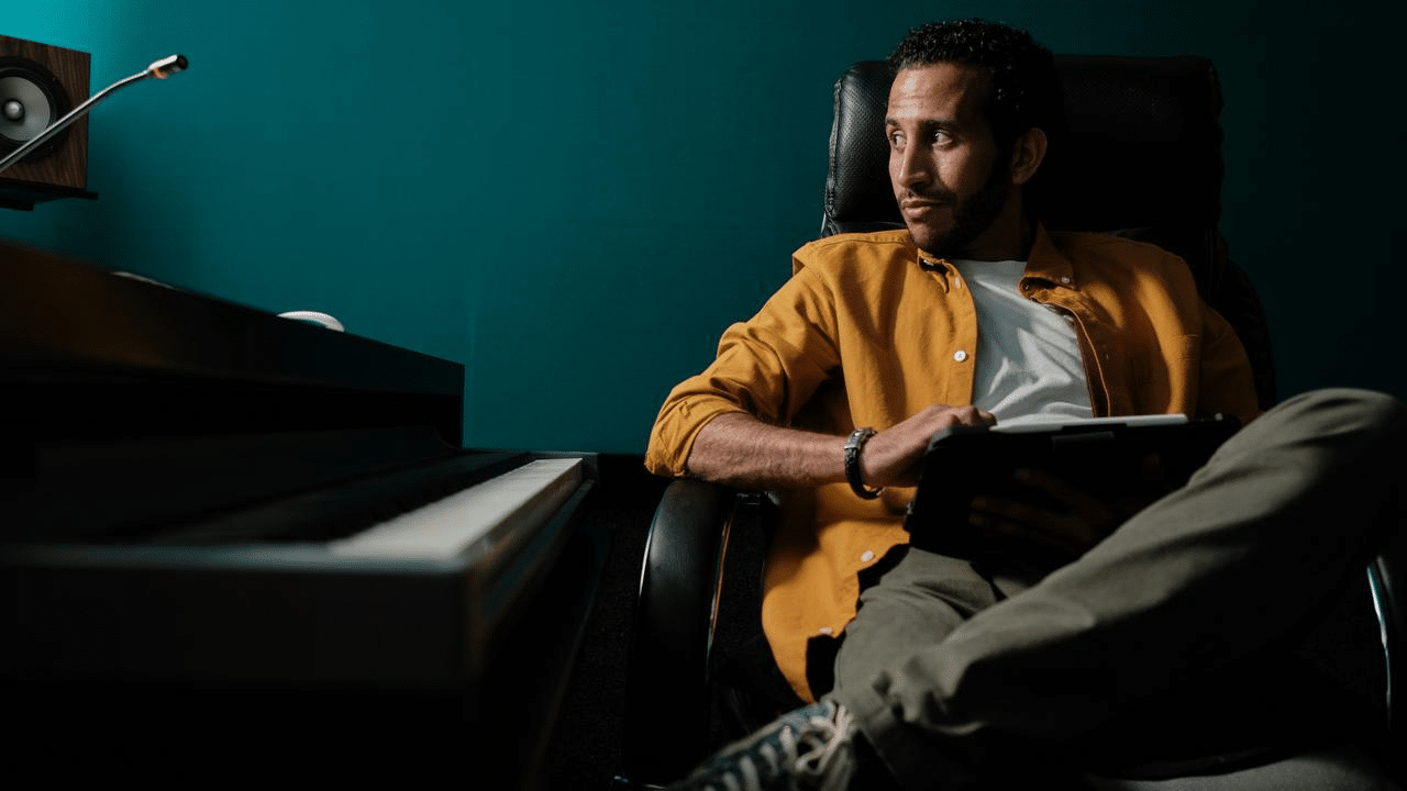 A man in a yellow shirt sitting in a chair holding a tablet near a piano.