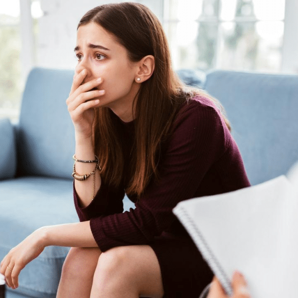 A young woman sitting on a couch looking distressed with her hand over her mouth.
