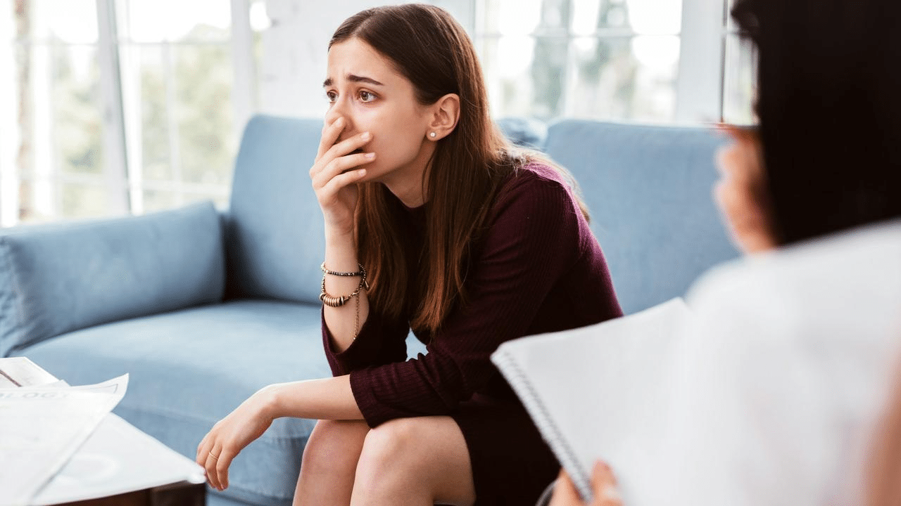 A young woman sitting on a couch looking distressed with her hand over her mouth.