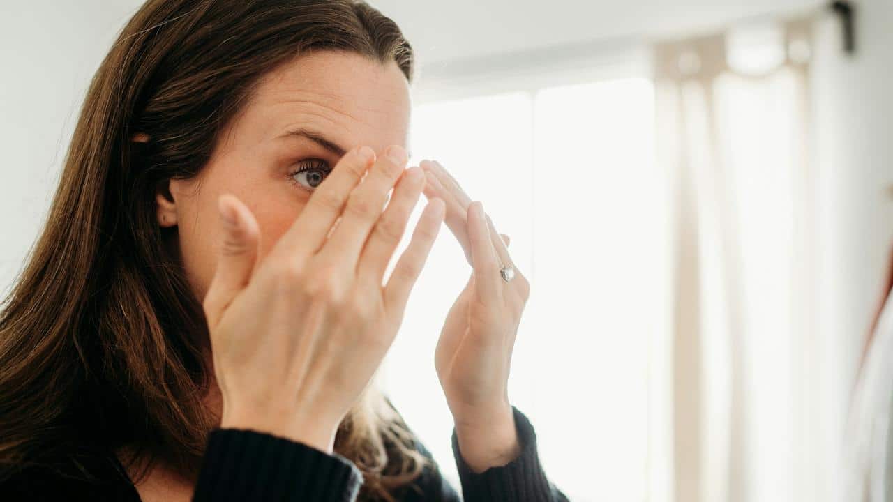 A woman applying skincare cream to her face in front of a mirror.