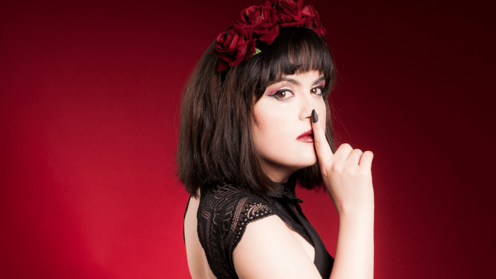 A woman in a lace top and a red rose headband holds a finger to her mouth.