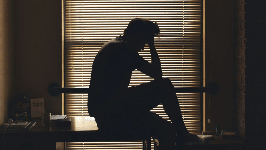 A silhouetted person sits on a desk with their head in their hands, with blinds in the background.