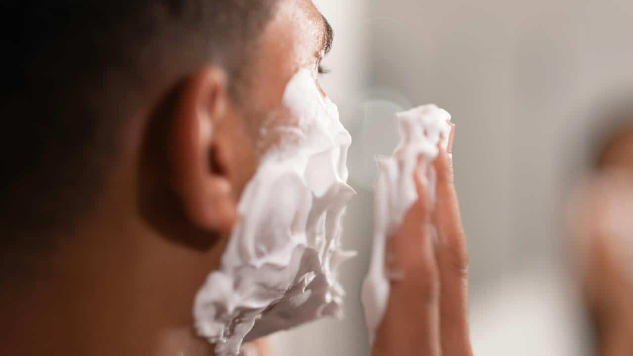 A man applying shaving cream to his face in front of a mirror.