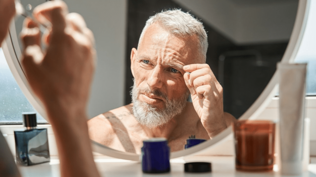 A man with gray hair and a beard looks at his reflection in a mirror, holding tweezers to his eyebrow.