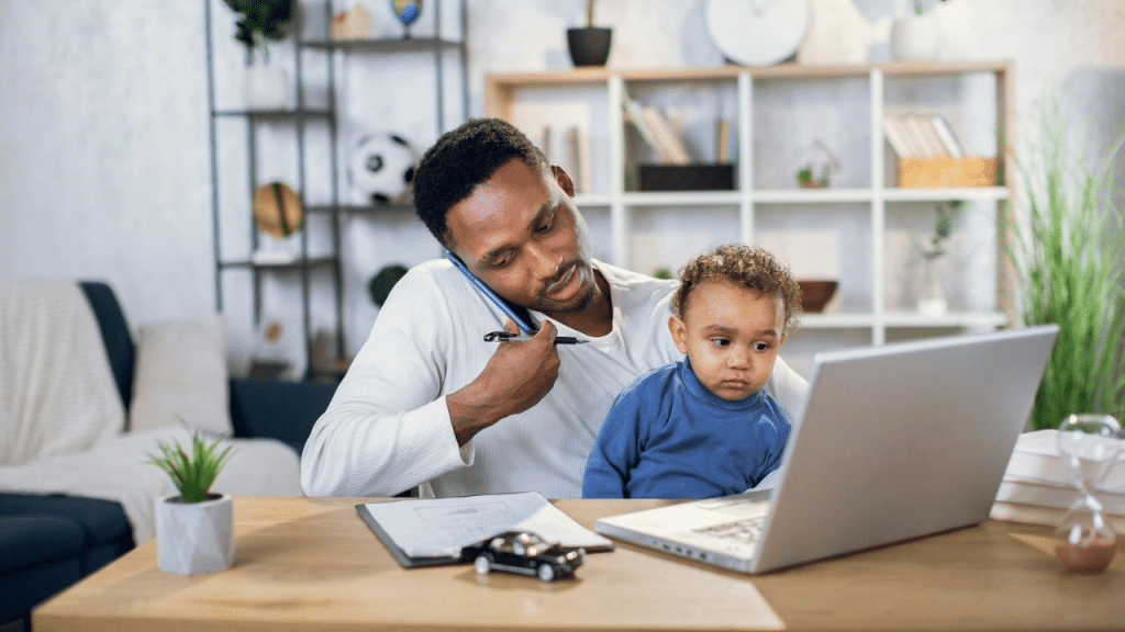A man sits at a desk, working on a laptop while holding a baby and talking on his phone.
