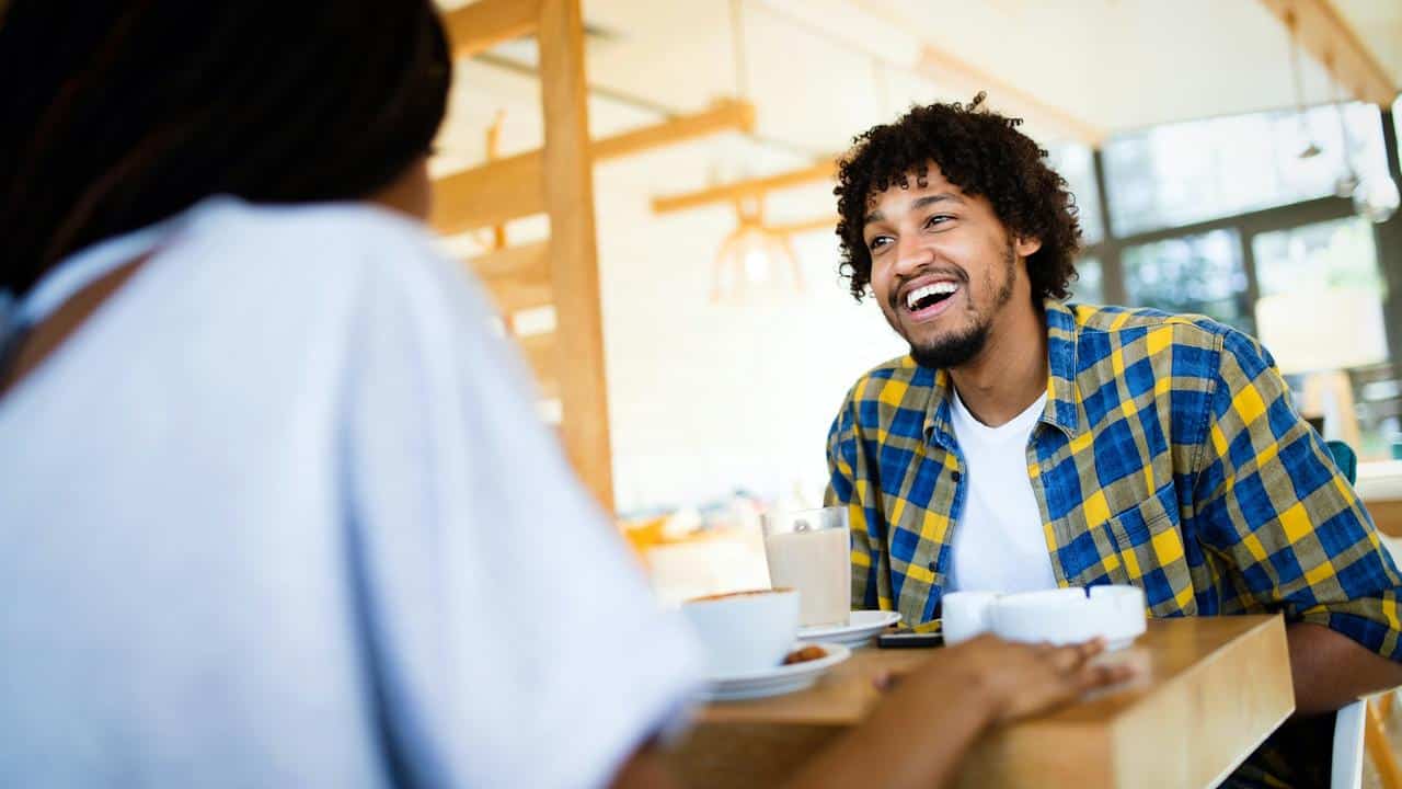 A man smiling while talking to a woman at a café table.