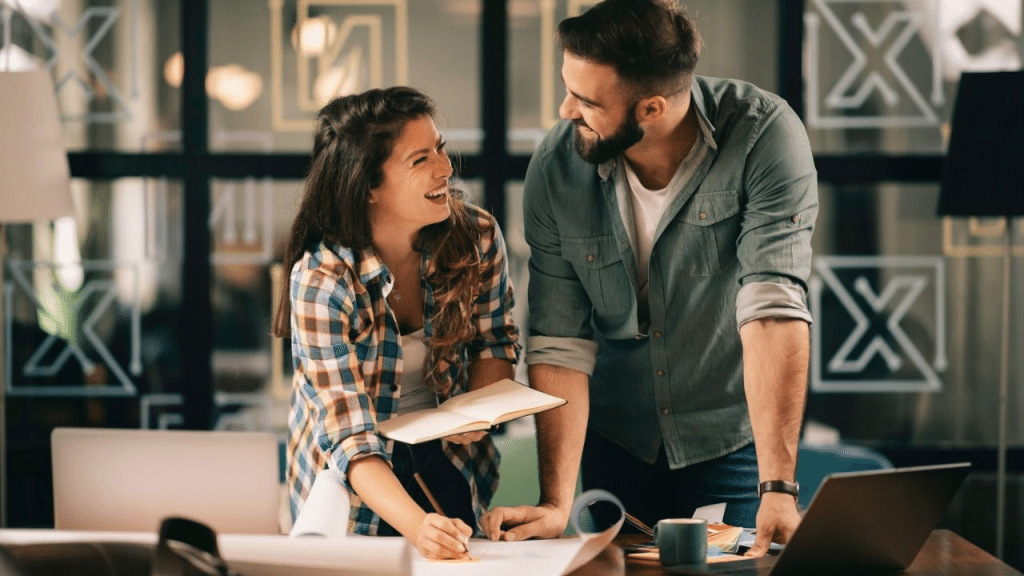 A happy man and woman lean over a desk together, smiling and looking at each other. The woman holds a notebook.