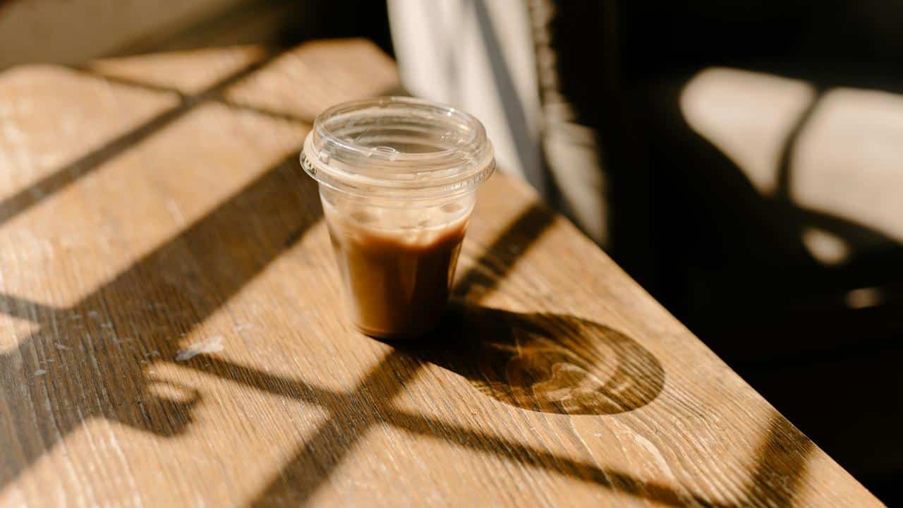 A plastic cup of iced coffee on a wooden table in sunlight.