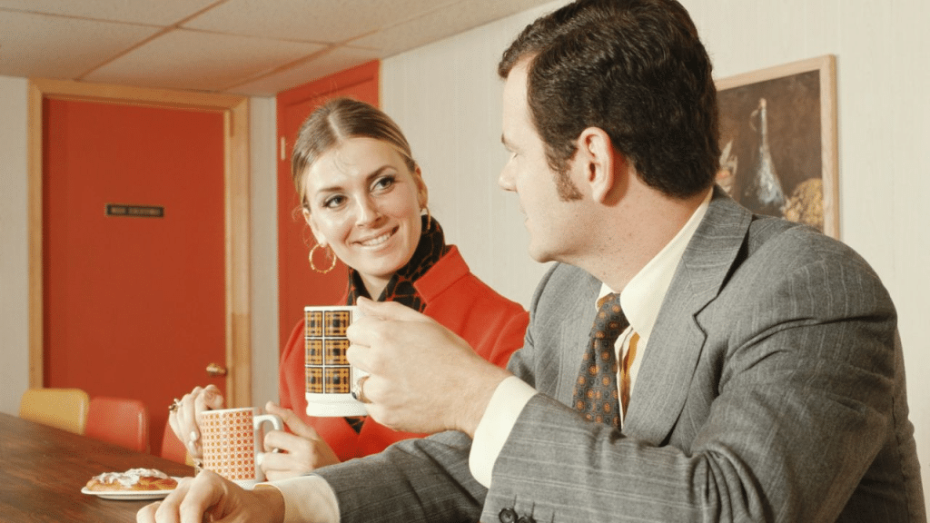 A smiling man and woman with a retro aesthetic sit at a table holding mugs.