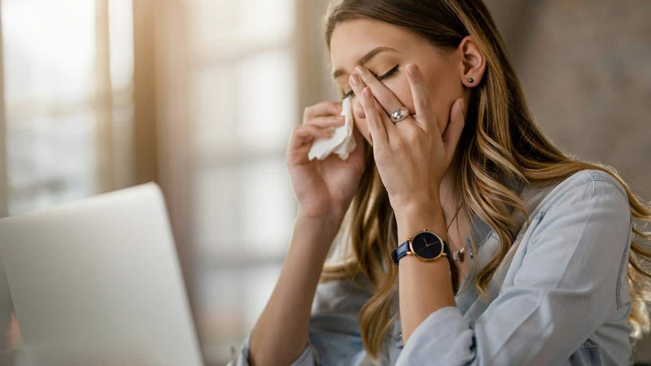 A woman sitting at a desk holding a tissue and rubbing her eyes.