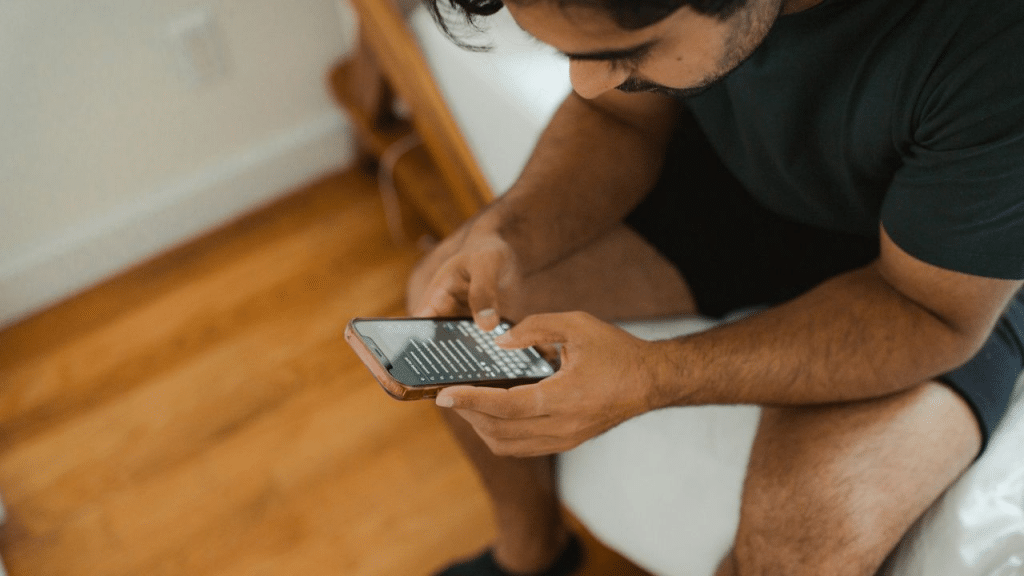 A man sits on the floor, looking intently at his phone with his thumbs on the screen.