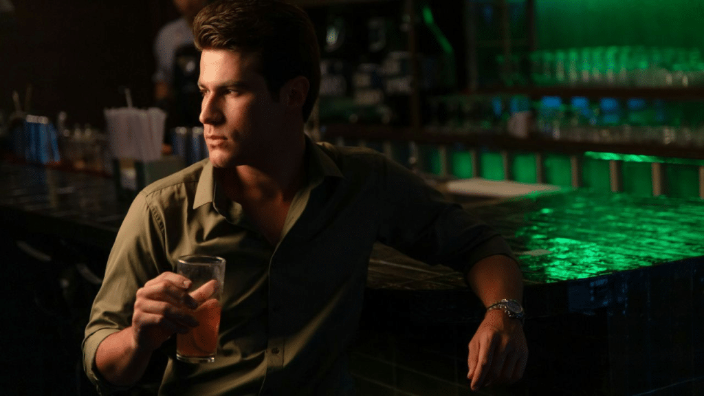 A man with dark hair leans on a bar counter, holding a drink and looking away.