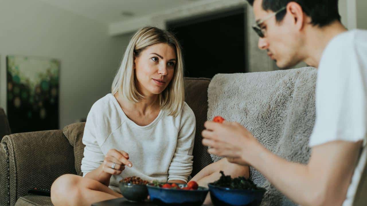 A woman and a man sitting on a couch having a serious conversation while eating snacks.