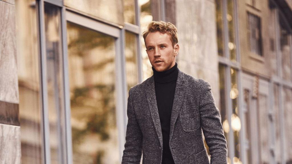 A man in a turtleneck and a gray blazer walks outside a building, looking off to the side.