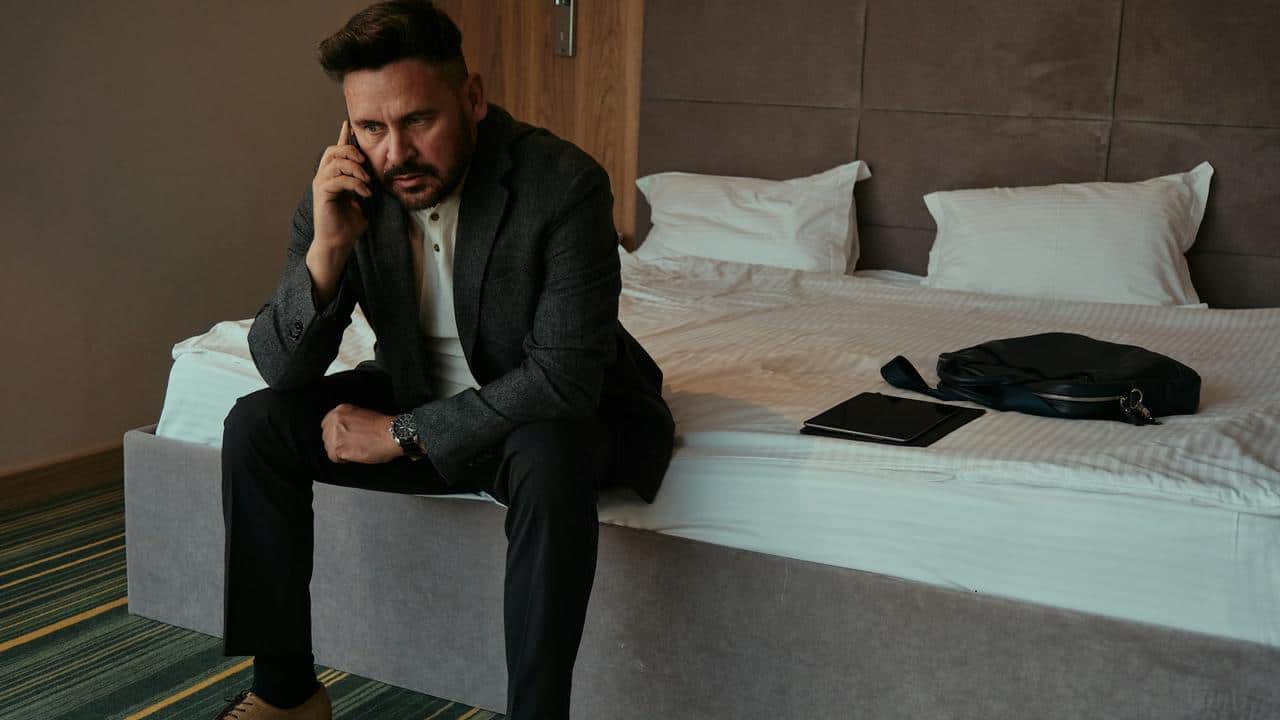 A man in a suit sitting on a bed while talking on the phone.