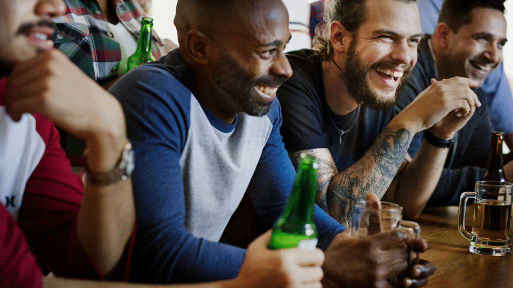 A diverse group of men smiling and laughing while drinking bottled beer at a bar.