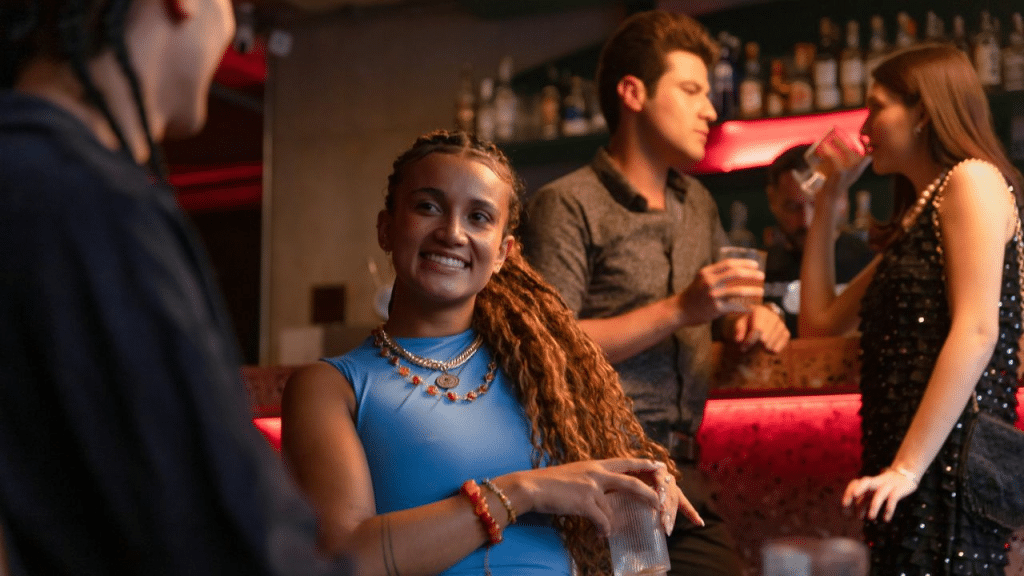 A young woman with long, braided hair smiles while holding a glass in a bar.