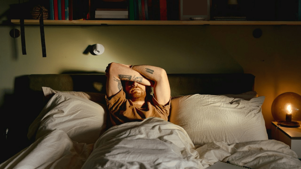 A man with tattoos on his arms is lying in bed, covering his eyes with his hands.