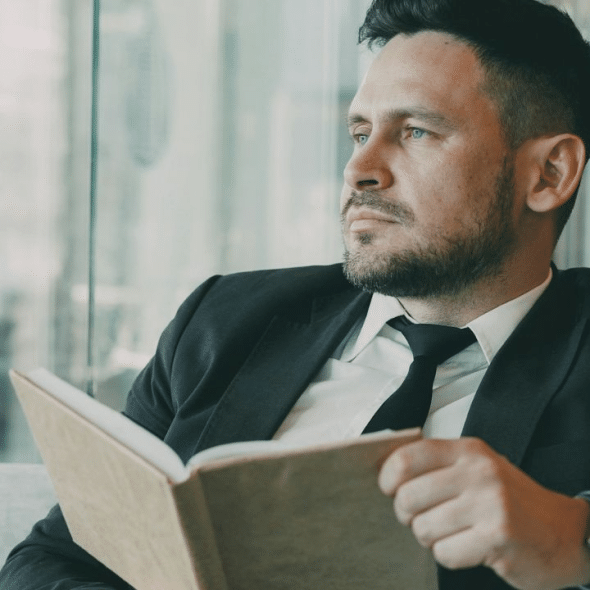 A businessman in a suit holds a book, looking thoughtfully out a window.