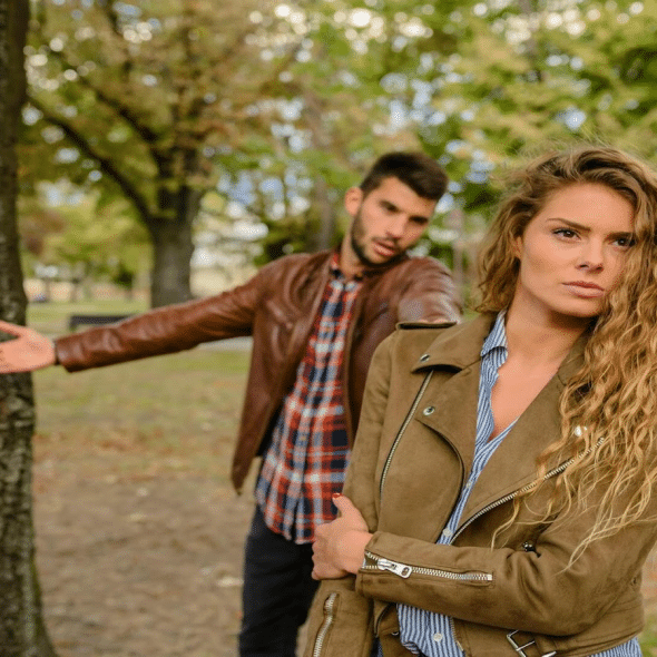 A serious woman with long curly hair turns away from a man gesturing with a questioning expression behind her in a park.
