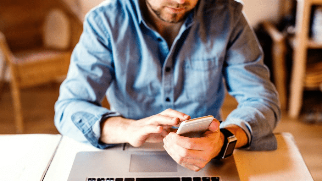 A man sitting at a desk using a smartphone with a laptop in front of him.