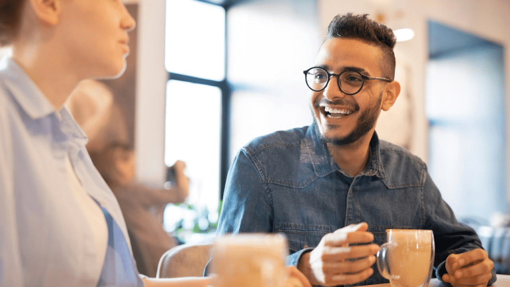 A man wearing glasses smiling while having coffee with a woman at a café.