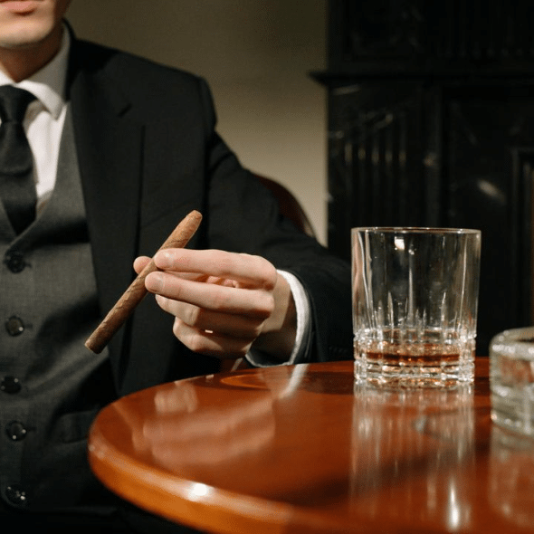 A man in a suit holding a cigar beside a glass of whiskey and a decanter.