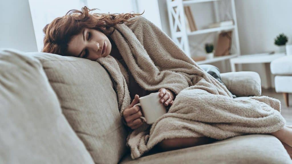 A woman rests on a couch wrapped in a blanket holding a mug.