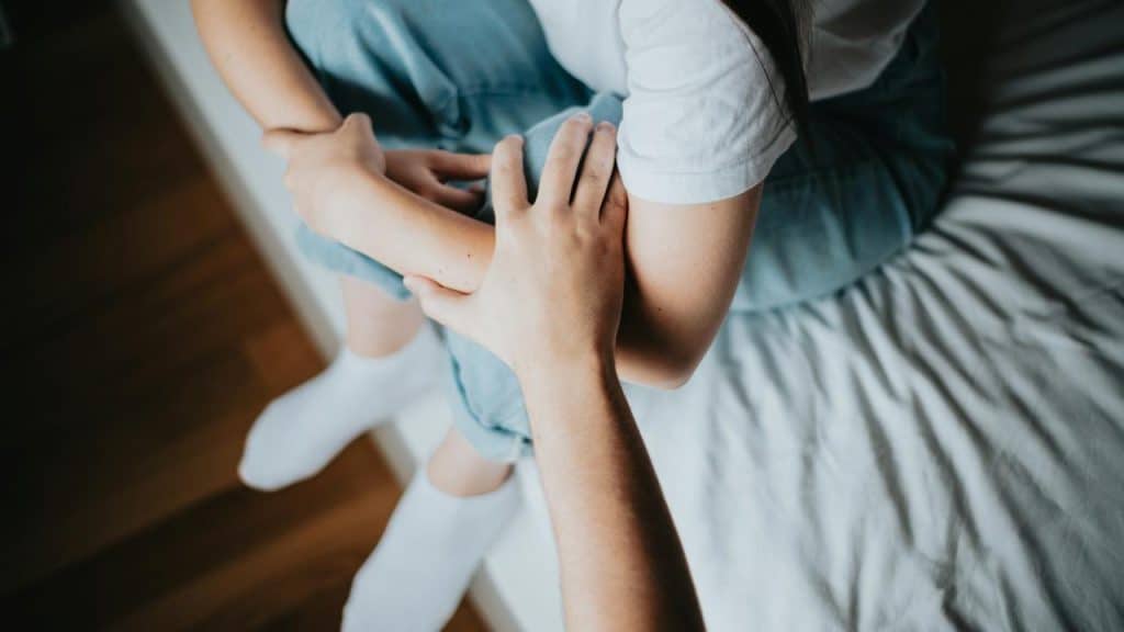 A person sits on a bed as another offers comfort with a hand on their arm.