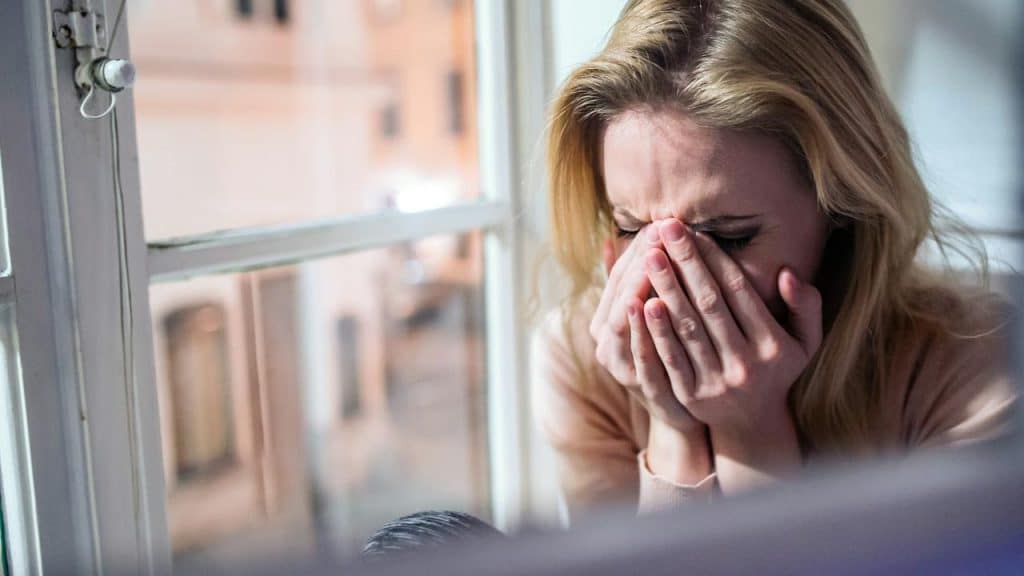 A woman cries with her face in her hands by a window.