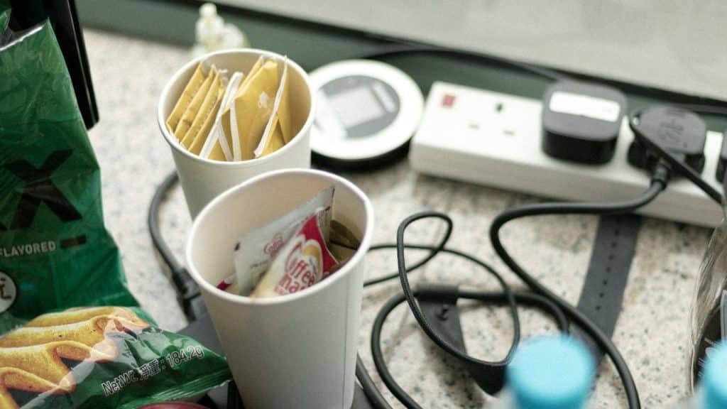 A cluttered table with snacks, cups, and power cords.