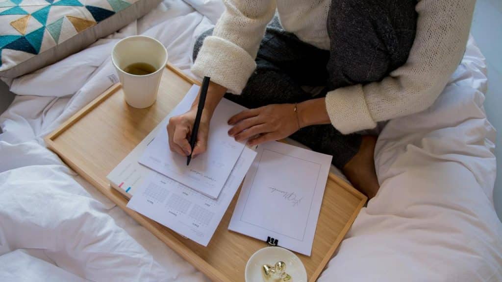 A person writes on papers with a cup of tea nearby on a bed tray.