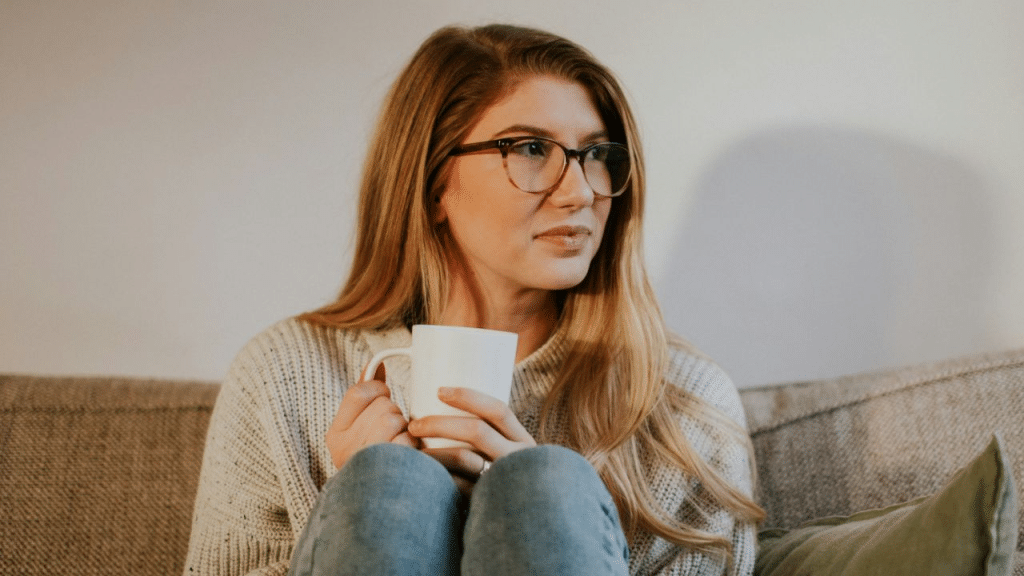 A thoughtful young woman in glasses and a sweater holds a mug on a couch.
