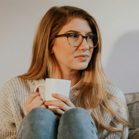 A thoughtful young woman in glasses and a sweater holds a mug on a couch.