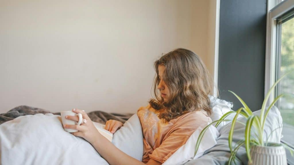 A woman reads a book while holding a mug in bed.