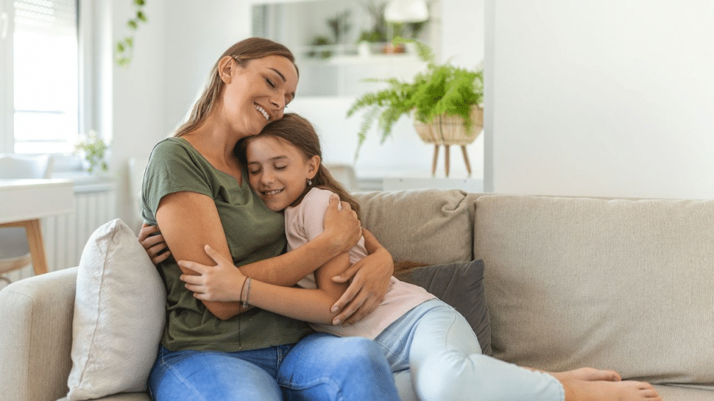 A mother in a green shirt and her young daughter in a pink shirt hug on a couch.