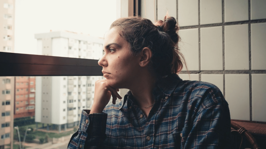 A young woman in a plaid shirt looks thoughtfully out of a window at city buildings.
