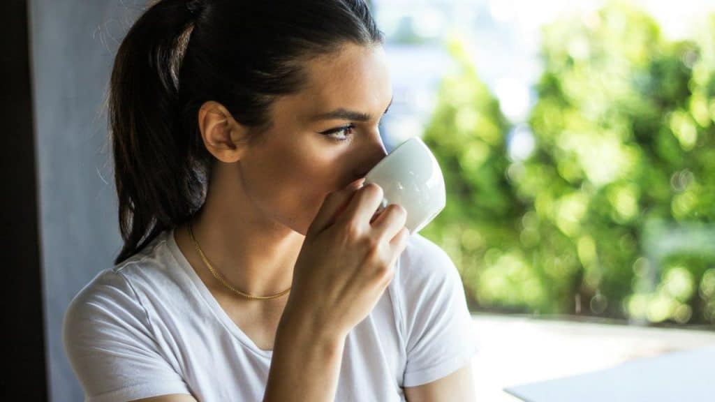 A woman drinks from a white mug while looking outside.