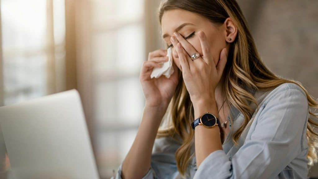 A woman wipes her eyes with a tissue while sitting at a laptop.