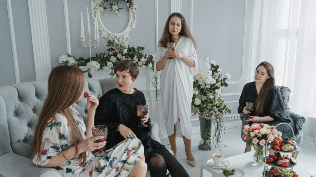 Four women in a formal living room are talking and drinking wine at a party.