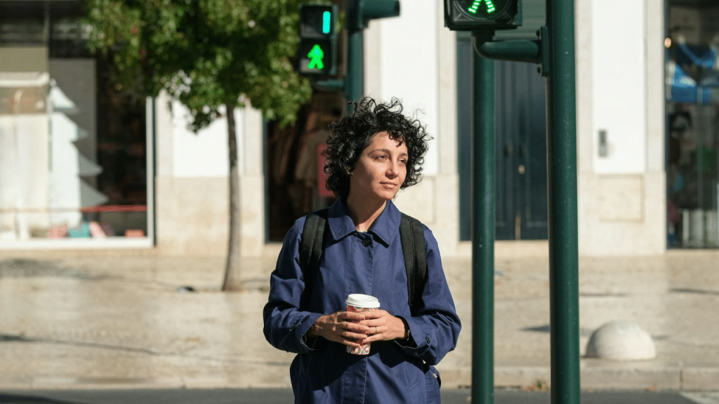A woman with curly hair stands at a street crosswalk, holding a coffee cup.